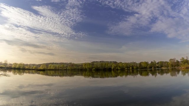 Timelapse of a lake over which clouds move with reflection on the water surface during the afternoon time until sunset with multi colored clouds near the town of Hustopece nad Becvou.