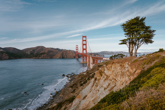 Horizontal photo of the famous Golden Gate Bridge. Rocky cliff above the ocean and green tree on the background of the bridge