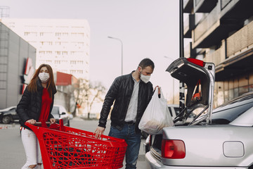 Couple in a city. Person in a mask. Coronavirus theme. People put shopping in the trunk.