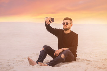 Fototapeta na wymiar Happy young bearded man in sunglasses taking a selfie on smartphone while sitting at the desert sand