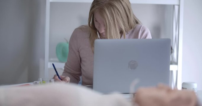 Serious blond Caucasian woman writing notes at the table with laptop as training mannequin for first aid learning lying at the foreground. Doctor getting ready for students exam. Cinema 4k ProRes HQ.