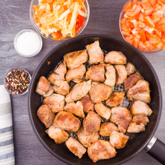 Fried pieces of pork in a pan and chopped vegetables in bowls close-up, top view