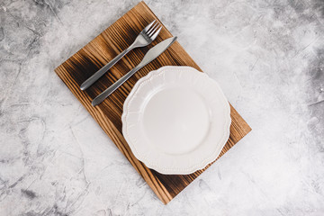 Empty white plate on a wooden board with cutlery on a gray background.	
