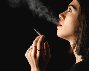 Fototapeta premium Low key close-up young woman smoking cigarette on black background. Copy space for text message.