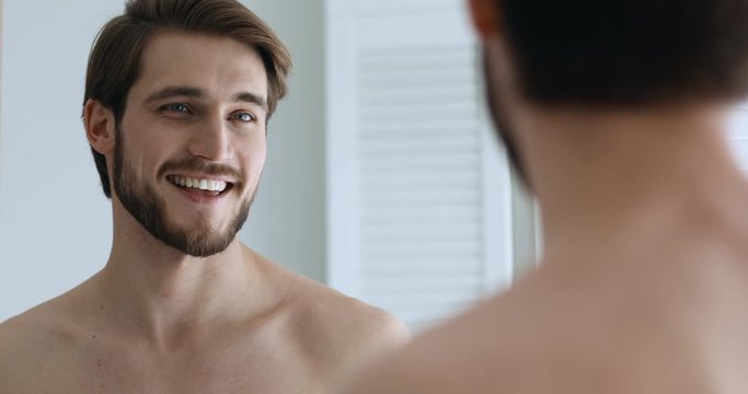 Funny confident young handsome man speaking to his reflection looking in bathroom mirror. Smiling sexy millennial guy narcissist touching beard feeling proud of himself shows good self-esteem concept.
