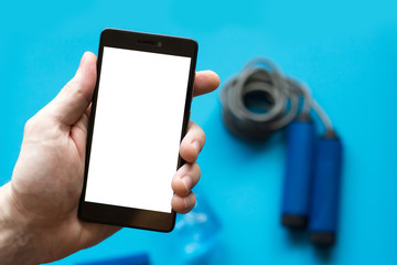 Smartphone with white screen in mans hand and sports equipment on blue background.