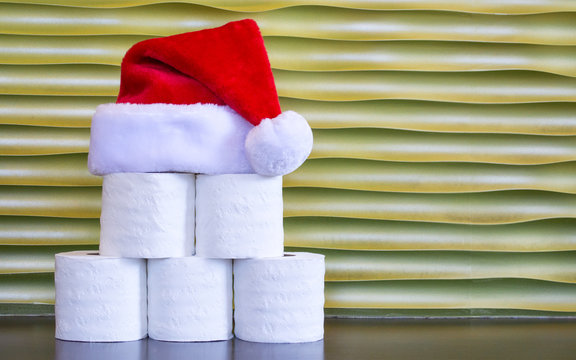 A pyramid of toilet paper with a red and white Santa Claus hat on top against a gold background with copy space