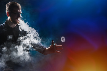 Fototapeta na wymiar Vaper in puffs of smoke. Man and smoke on a dark background. Ring from an electronic cigarette. Concept - buying a devas for a vaper. The guy is vaping. Components and accessories for vape.