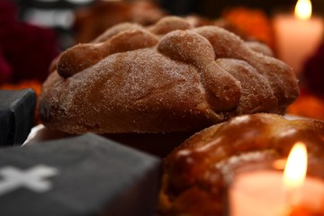 Pan de Muerto, traditional and sugared, around it marigold flowers, cardboard coffin, lighted candles, lit copal, diffused confetti background.