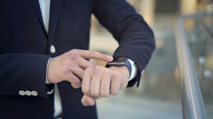 Close up of man dressed in business suit white shirt and blue jacket using smartwatch on his left hand. On background of blurred floor in shopping center