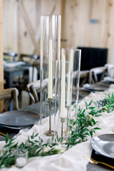 Cylinder Candle Holders with Tall Taper Candles Decorate a Table at a Wedding Reception, Decorated with Olive Leaves on Loose Tablecloth Cheesecloth Runner. 