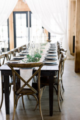 Wedding Reception Table with Antique Chairs with a Dinner Table with Gray Plates, Greenery, and Tall Glass Candleholders and Taper Candles. 