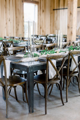 Close-up at wedding reception table design. Criss-crossed antique brown chairs at gray long dinner tables decorated with gray plates, green candle garland, and tall taper candles. 