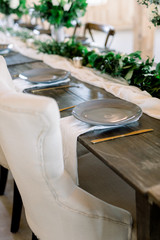 Fototapeta na wymiar Gray plates with gold silverware on a wood table with a runner made of green eucalyptus and olive leaves at a wedding reception. 