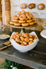 A bowl of delicious donut holes on a Wood Table. In the background, a cake stand piled with glazed donuts sits. There is a donut wall chart in the background. 