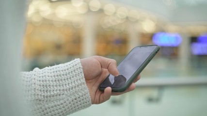 Close up of man s hand in white sweatshirt scrolling smartphone on blurred yellow background. Left male hand with a smartphone indoors on yellow bokeh