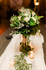 Flower Bouquet with White Roses, Baby's Breath and Eucalyptus Leaves in a Vase at the Head Table at a Wedding with Votive Candles and a White Cheesecloth on a Wood Table