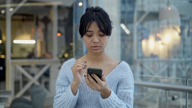 Portrait close up handheld shot of smiling happy black hair asian female standing and holding scrolling a phone on background of hitech office