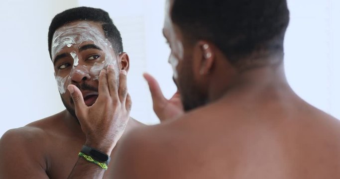 Handsome young adult afro american man applying facial mask cream on face looking in mirror. African shirtless guy doing morning skin care beauty routine. Male skincare dermatology treatment concept.