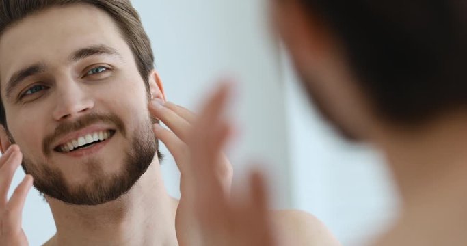 Handsome confident smiling young adult unshaven man touching grooming beard in the morning. Happy charming groomed guy with bristle looking in bathroom mirror. Men beauty concept. Close up view