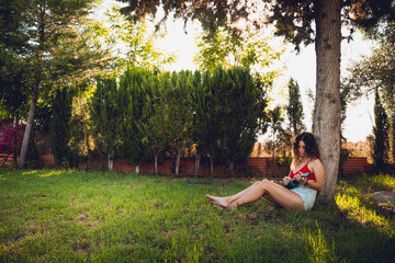 Naklejka na ściany i meble Young adult woman wearing a red bikini is playing a ukelele sitting under a tree in a park. She is focus while playing it.