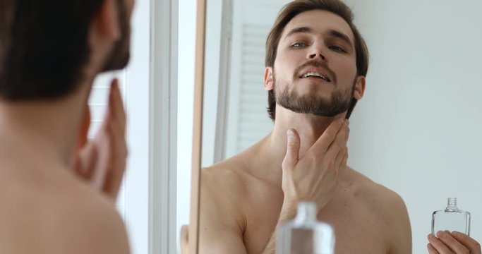 Smiling confident handsome young guy applying aftershave perfume lotion on face. Happy sexy shirtless bearded man putting fragrance on neck skin after shaving looking in mirror. Men grooming concept.