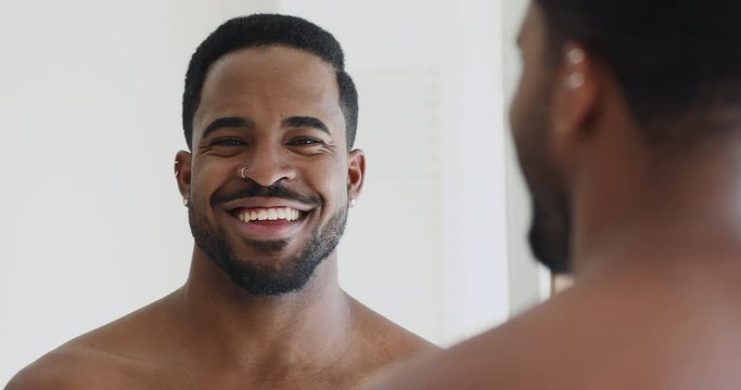 Happy handsome afro american ethnic young adult hipster man with piercing looking in mirror at camera. Attractive shirtless millennial african guy with healthy teeth, dental smile, close up portrait.