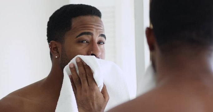 Happy fresh african young hipster man wiping face with towel looking in bathroom mirror. Smiling sexy shirtless afro american guy doing washing, shaving, grooming morning hygiene routine concept.