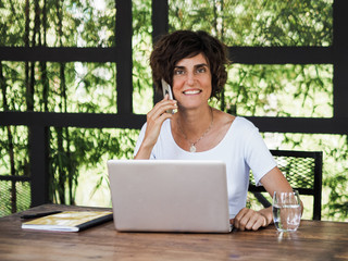 happy smiling remote digital nomad woman talking on the mobile phone with laptop notebook, pen and glass in white shirt sitting at a work desk in a co-working cafe green plants and black structures