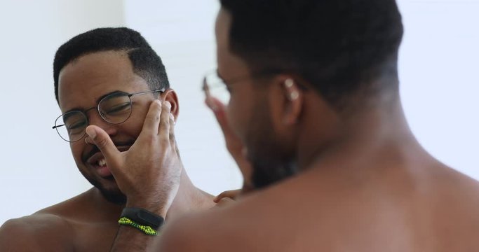 Happy attractive young adult african man wearing glasses grooming looking in bathroom mirror. Confident bearded afro american metrosexual hipster guy getting ready, doing morning beauty routine.