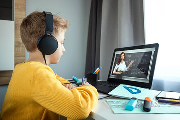 Distance learning, a boy in headphones sits at a table at home looking in a beech laptop. The...