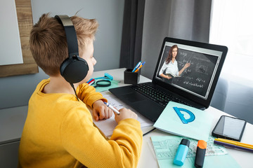 Distance learning, a boy in headphones sits at a table at home looking in a beech laptop. The concept of online education, home education, technology, school.