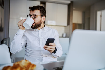 Fototapeta na wymiar Young cheerful handsome bearded man sitting at dinning table at home and drinking fresh morning coffee. In other hand he is holding smart phone.