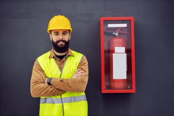 Young smiling cheerful attractive bearded worker in vest, with helmet on head standing next to fire...