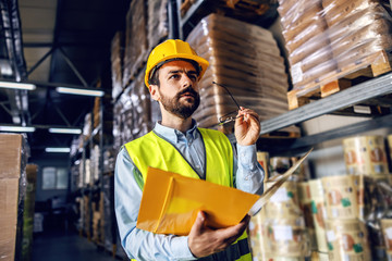 Young serious bearded supervisor in vest holding folder with documents and checking out goods in warehouse.