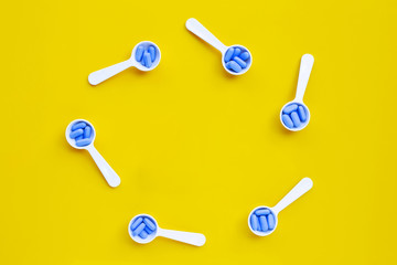 Frame made of medical blue pills with white plastic spoon on yellow background.