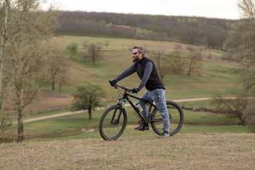 Fototapeta premium Sports brutal bearded guy on a modern mountain bike. Cyclist on the green hills in the spring.
