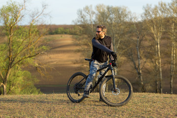 Fototapeta premium Sports brutal bearded guy on a modern mountain bike. Cyclist on the green hills in the spring.