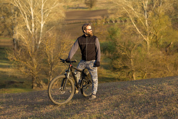 Sports brutal bearded guy on a modern mountain bike. Cyclist on the green hills in the spring.