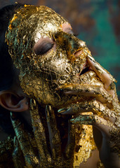 Girl with a mask on her face made of gold leaf. Gloomy studio portrait of a brunette on an abstract background.