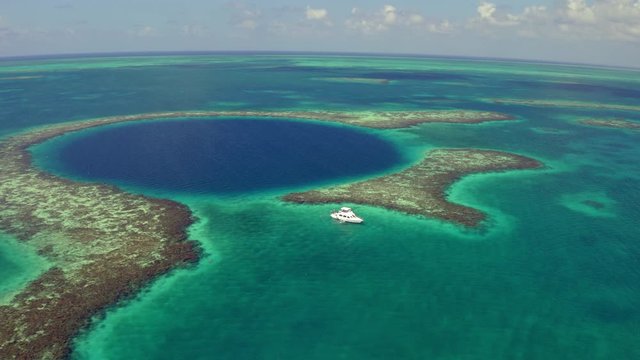 Aerial view of yacht next to large marine sinkhole on sunny day, drone panning from left to right over sea against sky - Great Blue Hole, Belize