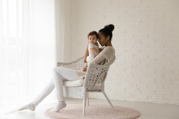Loving young biracial mother sit in chair in living room hold hug small newborn baby relax at home...