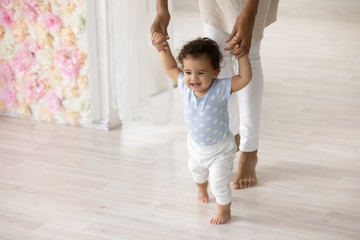 Smiling little biracial toddler infant child make first steps at home holding mom hands, happy small african American baby learn walking with mother support and care, upbringing, childcare concept - Powered by Adobe