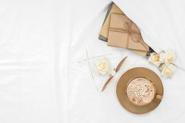 Correspondence letters, flowers, book, coffee cup with foam, reading glasses on the background of a crumpled white bedsheet. Cozy waking in bed Top view. Flat lay