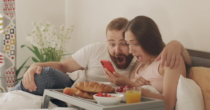 Young man and woman looking at phone screen and having good news while lying on bed. Happy couple using smartphone with amazed face and saying woow during breakfast in bedroom.