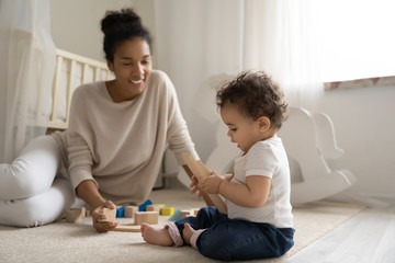 Happy young biracial mother sit on floor play with building blocks bricks with little infant...