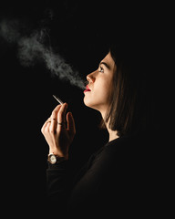 Fototapeta premium Low key close-up young woman smoking cigarette on black background. Copy space for text message.