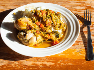 portion of Chakhokhbili (traditional georgian dish from stewed chicken, tomato, eggplant and fresh herbs) with boiled rice on white plate on wooden table in home kitchen illuminated by sun
