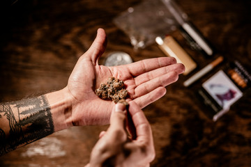 A hand holding compact marihuana buds with tobacco, lighter and grinder in the background. This dark mood layout matches with the fact of being illegal substance in most countries. Concept: Marihuana