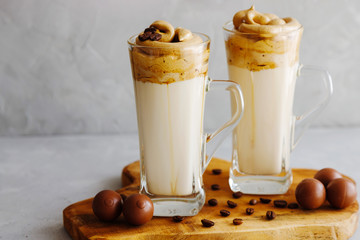 Two glasses of Dalgon stand on a wooden board and next to them are chocolates. Trendy Milk Drink with Coffee Sweet Foam